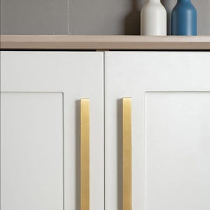 May include: White cabinet doors with vertical gold-coloured handles. Two decorative bottles, one white and one blue, sit on a shelf above the doors. The background is a light grey wall.
