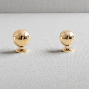 Peut inclure: Deux boutons de meuble sphériques dorés. Chaque bouton a une surface polie et réfléchissante, reposant sur une base cylindrique courte. Ces boutons sont probablement destinés aux meubles ou à la décoration.