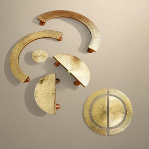 Może przedstawiać: Zestaw mosiężnych okuć do szafek w różnych kształtach, w tym łukowate uchwyty, okrągła gałka i półokrągłe uchwyty. Okucia mają teksturowaną powierzchnię i drewniane akcenty.