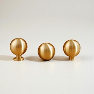 May include: Three gold-coloured cabinet knobs with a spherical design. One knob has a small base, while the other two have a shorter, cylindrical base. The knobs are made of a shiny, metallic material.