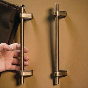 May include: Two antique brass cabinet pulls with a rectangular base and a vertical bar. The pulls are mounted on a tan surface. One pull is being held by a hand. The word "NUVANA" is in the upper left corner.