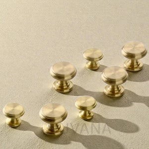 Op de afbeelding: Een verzameling messing kastknoppen met een ronde boven- en onderkant en een cilindrische steel. De knoppen zijn gerangschikt op een neutrale ondergrond en werpen schaduwen. De hardware heeft een gepolijste, metallic afwerking.