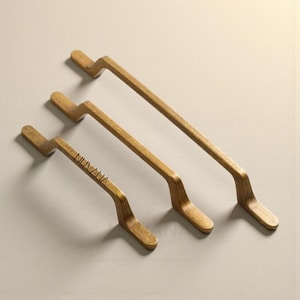 May include: Three brass-toned cabinet pulls with a modern, angular design. The pulls are arranged diagonally, showcasing their varying lengths. The smallest pull has the word "NUVANA" printed on it. The hardware is set against a neutral background.