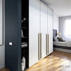 May include: Modern bedroom wardrobe with a navy blue side and white doors. The wardrobe has gold-colored handles and an open shelf with decorative items. The room features a bed with pillows and a wooden floor.