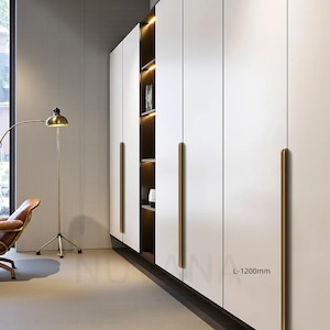 Könnte beinhalten: Ein moderner weißer Kleiderschrank mit langen, goldfarbenen Griffen. Der Schrank verfügt über ein eingebautes Regal mit warmer Beleuchtung. Das Design ist schlicht und minimalistisch, passend für moderne Wohnkultur.