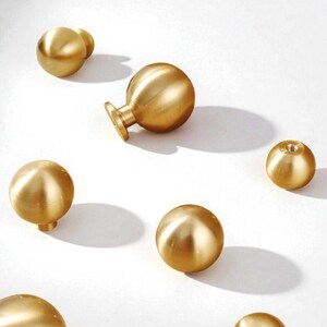 May include: Several brass-finish cabinet knobs in different shapes, including spherical and mushroom designs. The knobs are displayed on a white surface, highlighting their metallic lustre and simple elegance. Suitable for furniture upgrades.