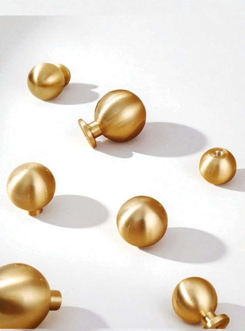 May include: Several brass-finish cabinet knobs in different shapes, including spherical and mushroom designs. The knobs are displayed on a white surface, highlighting their metallic lustre and simple elegance. Suitable for furniture upgrades.