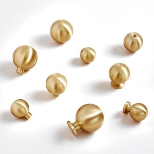 May include: An assortment of brass-coloured cabinet knobs in different sizes and styles. The knobs are spherical, some with a stem or flat base. The warm gold tone and simple shapes suggest a modern or vintage aesthetic. Measurements are in centimetres.