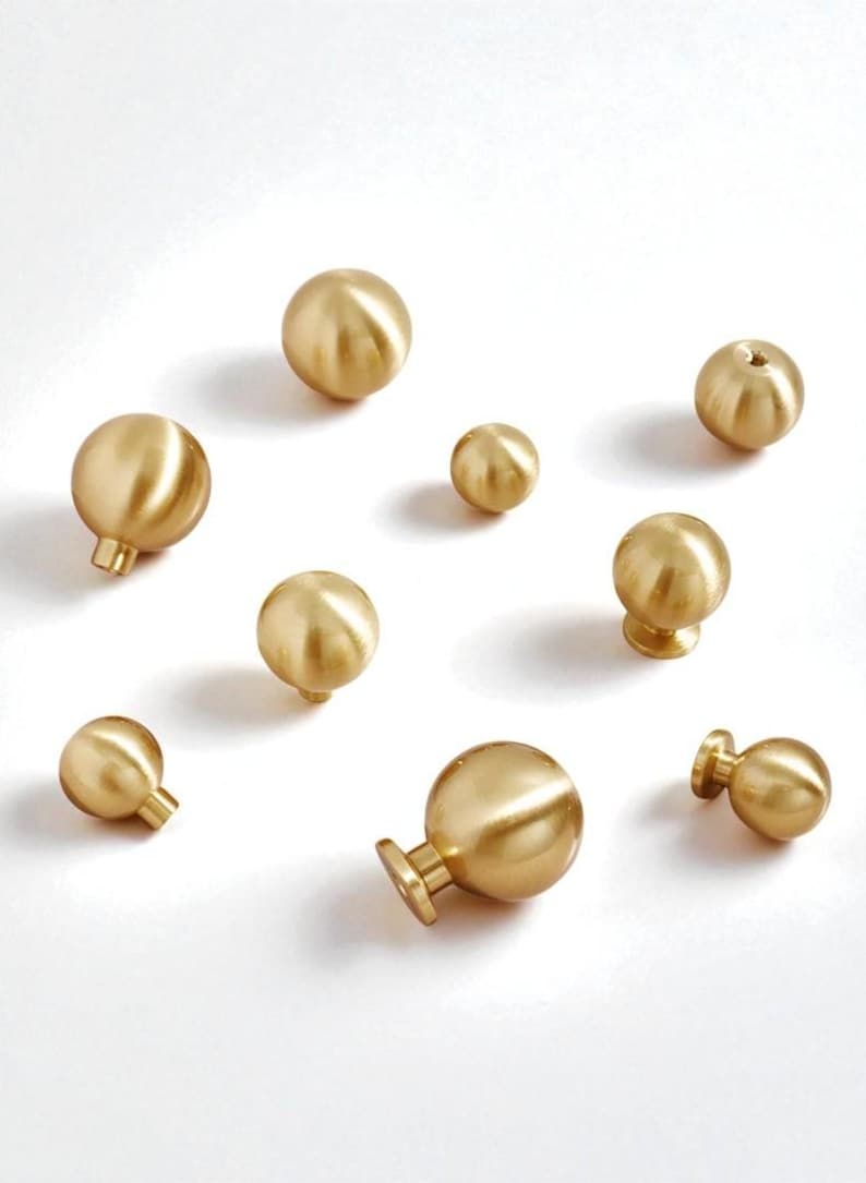 May include: An assortment of brass-coloured cabinet knobs in different sizes and styles. The knobs are spherical, some with a stem or flat base. The warm gold tone and simple shapes suggest a modern or vintage aesthetic. Measurements are in centimetres.