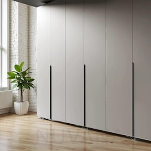 Op de afbeelding: Een rij lichtgrijze, moderne garderobedeuren met zwarte verticale handgrepen. De deuren staan tegen een witte bakstenen muur en een lichte houten vloer. Een potplant staat in de hoek.