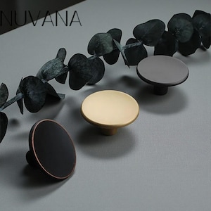 Peut inclure: Quatre boutons de meubles ronds, de couleurs variées : noir avec un bord cuivré, doré et gris foncé. Les boutons ont un dessus plat et une base cylindrique courte. Le mot "NUVANA" est visible en haut à gauche.