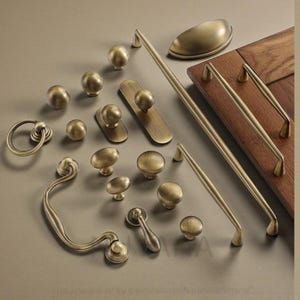 Könnte beinhalten: Eine Auswahl an antiken Messing-Schrankbeschlägen, darunter Knöpfe, Griffe und Ziehgriffe. Die Kollektion umfasst verschiedene Formen und Größen, von runden Knöpfen bis zu länglichen Ziehgriffen, alle in einem warmen, gealterten Messing-Finish. Eine Holztür ist im Hintergrund zu sehen.