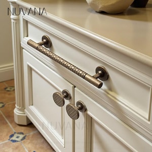 May include: A white cabinet with bronze handles and knobs. The long handle has a textured design, and the knobs feature a honeycomb pattern. The word "NUVANA" is visible on the cabinet. The cabinet is a piece of furniture.