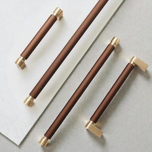 Peut inclure: Ensemble de poignées de meubles marron et dorées. Les poignées présentent une texture côtelée et des embouts dorés. Le matériel est disposé en diagonale sur une surface gris clair.