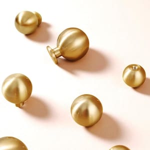 May include: A collection of gold-coloured, spherical cabinet knobs are arranged on a pale pink surface. The knobs have a smooth, metallic finish and vary in design, with some featuring a small stem or base for attachment. The overall aesthetic is modern and minimalist.