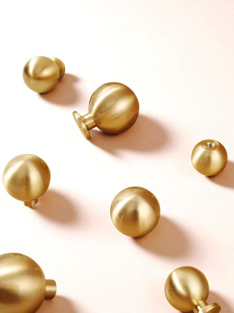 May include: A collection of gold-coloured, spherical cabinet knobs are arranged on a pale pink surface. The knobs have a smooth, metallic finish and vary in design, with some featuring a small stem or base for attachment. The overall aesthetic is modern and minimalist.