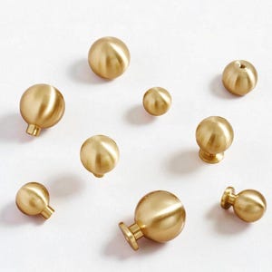 May include: A selection of brass cabinet knobs in different sizes, displayed on a white background. The knobs are spherical with a polished, gold finish. Some have a stem and base for mounting.