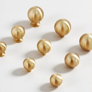 May include: A selection of gold-coloured, spherical cabinet knobs. The knobs have a smooth, metallic finish and vary in size. Some have a small, cylindrical base, while others have a flared base. The knobs are arranged on a white surface, creating a clean aesthetic.
