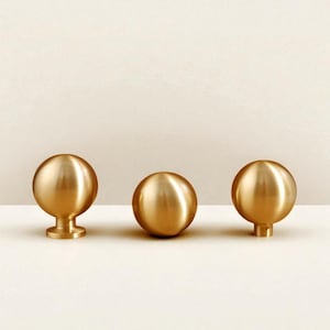 May include: Three polished brass cabinet knobs. Two have a spherical design with a small base, and one has a cylindrical base. The knobs are arranged in a row against a neutral background. Ideal for drawers or cupboards.