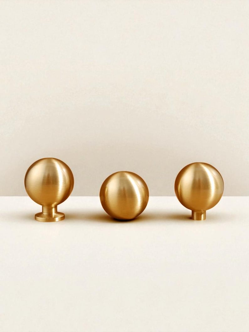 May include: Three polished brass cabinet knobs. Two have a spherical design with a small base, and one has a cylindrical base. The knobs are arranged in a row against a neutral background. Ideal for drawers or cupboards.