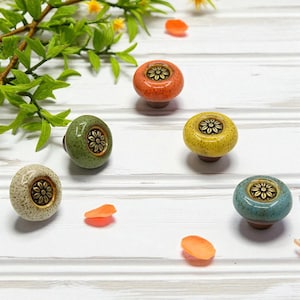 May include: A collection of five colorful ceramic cabinet knobs. Each knob features a floral design in the center and is in shades of orange, green, yellow, blue, and cream. The knobs are set against a white wooden background.