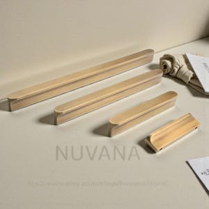 Puede incluir: Cuatro tiradores de madera para armarios de diferentes tamaños, dispuestos sobre una superficie clara. Los tiradores tienen forma rectangular con extremos redondeados y un acabado de madera natural. La palabra "NUVANA" está impresa en la superficie.