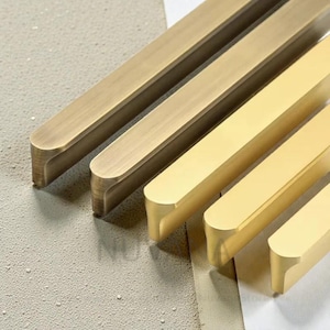 May include: Close-up of several brass cabinet pulls in varying sizes. The pulls have a rectangular shape with rounded ends and a brushed finish. The brass colour ranges from a darker, antique tone to a brighter, polished gold.
