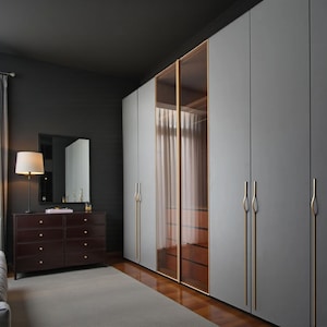 May include: A modern bedroom interior featuring a large gray wardrobe with gold handles and glass-paneled doors. A dark brown dresser with a mirror and lamp sits to the left. The room has a gray carpet and dark walls.