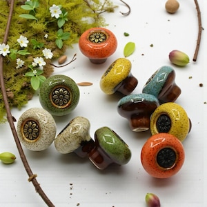 Puede incluir: Una colección de coloridos tiradores de cerámica en varios tonos de naranja, verde, amarillo y azul. Cada tirador presenta un diseño floral decorativo en el centro. Los tiradores se exhiben sobre una superficie blanca con elementos naturales.