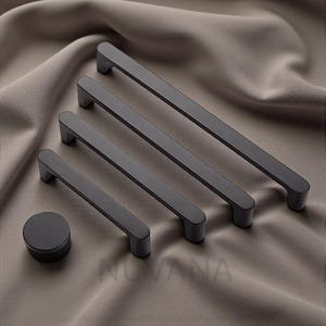 May include: A set of four matte black cabinet pulls and a small round knob. The pulls are rectangular with rounded ends, varying in length. The hardware is displayed on a neutral-toned fabric.