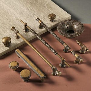 Pode incluir: Uma variedade de ferragens de armário de latão antigo, incluindo puxadores, puxadores e uma tigela decorativa. As ferragens apresentam vários designs, como superfícies texturizadas e padrões lineares. As peças são dispostas sobre uma superfície de madeira clara e um fundo marrom avermelhado.