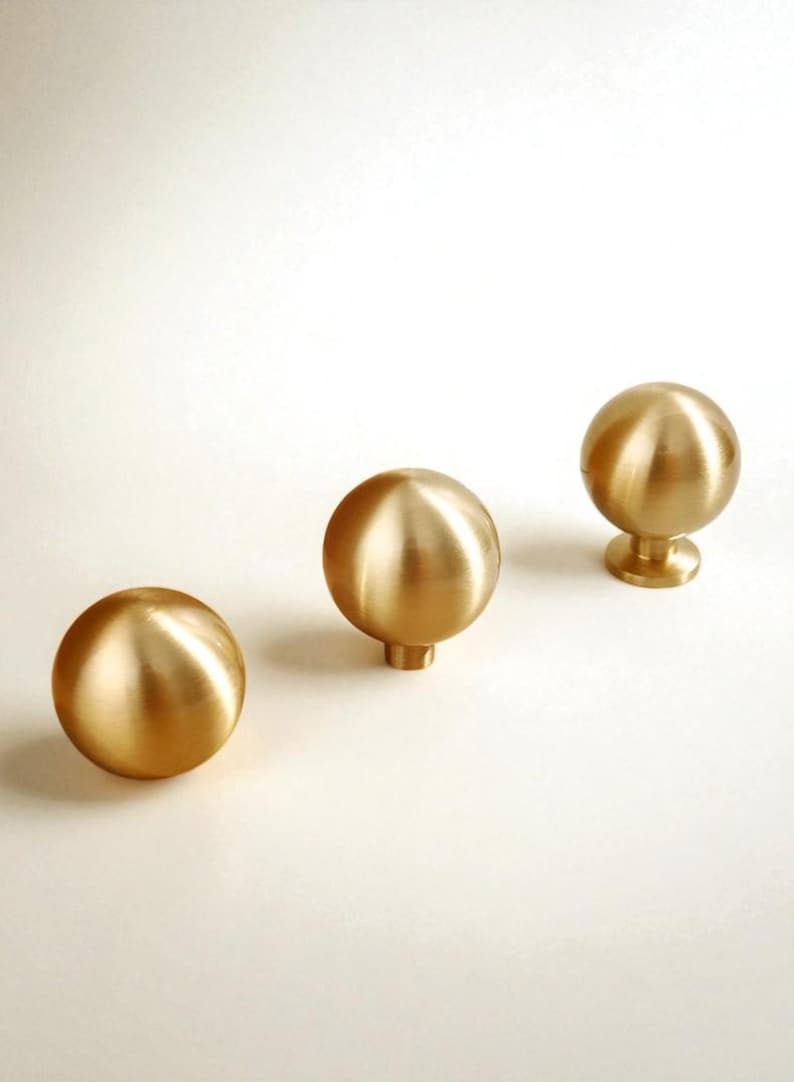 May include: Three brass cabinet knobs with a spherical design. The knobs have a polished finish and are arranged on a white surface. The knobs are approximately 3.8 cm in diameter.