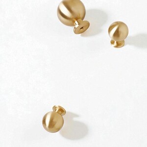 May include: Three gold-coloured, spherical cabinet knobs with a brushed finish. Each knob has a short, cylindrical base with a screw hole. The knobs are arranged on a white background, with varying shadows.