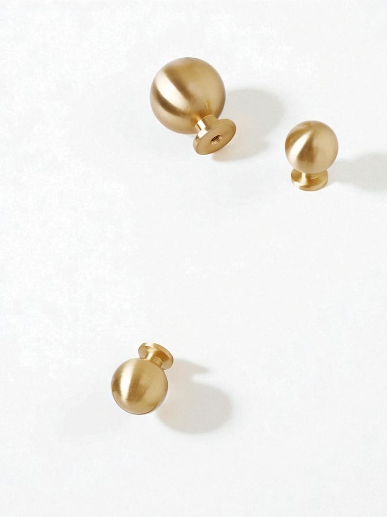 May include: Three gold-coloured, spherical cabinet knobs with a brushed finish. Each knob has a short, cylindrical base with a screw hole. The knobs are arranged on a white background, with varying shadows.