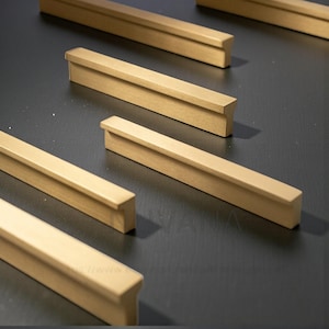 Peut inclure: Gros plan de plusieurs poignées de meubles dorées disposées sur une surface sombre. Les poignées ont une forme rectangulaire avec un bord légèrement surélevé d'un côté. L'éclairage met en valeur la finition métallique et les lignes épurées.