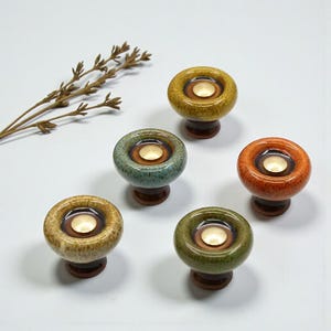 May include: Five ceramic cabinet knobs in shades of yellow, green, and orange. Each knob has a circular design with a light-colored center and a dark brown base. The knobs are arranged on a white surface.
