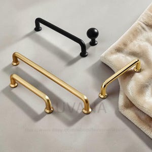 May include: An assortment of cabinet hardware in matte black and gold finishes. The selection includes bar pulls and a round knob. The gold hardware is in various sizes. The hardware is displayed on a neutral surface.