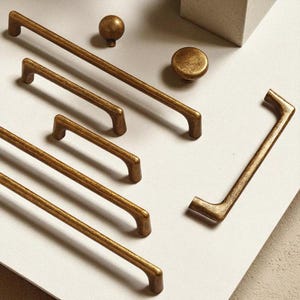 May include: An assortment of antique brass cabinet hardware, including various sizes of bar pulls, a knob, and a round pull. The hardware has a distressed finish, giving it a vintage look. The pieces are arranged on a neutral surface.