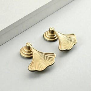 Puede incluir: Dos tiradores de armario dorados con un diseño en forma de abanico. Los tiradores son de metal y tienen una superficie detallada y texturizada. La ferretería está sobre un fondo gris claro.