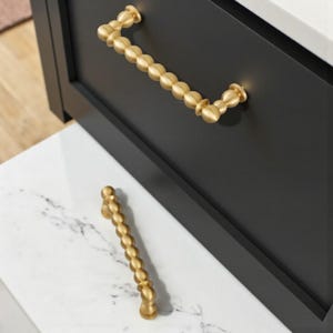 Pode incluir: Ferragens de armário douradas com design de contas. A puxador da gaveta está fixada a um armário preto. Um segundo puxador repousa sobre uma superfície de mármore branco e cinza. A ferragem tem um acabamento metálico quente.