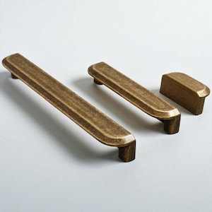 Könnte beinhalten: Drei antike Messing-Schrankgriffe und ein Knauf. Die Griffe sind rechteckig mit abgerundeten Kanten und einer strukturierten Oberfläche. Der Knauf hat eine kleine, rechteckige Form. Alle Teile haben eine warme, gealterte Oberfläche.