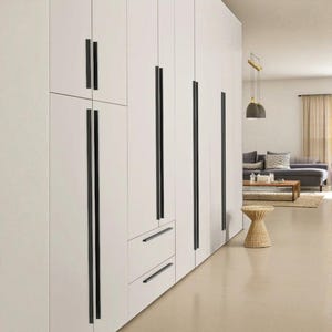 Könnte beinhalten: Ein weißes Einbauschranksystem mit schwarzen vertikalen Griffen. Die Schranktüren und Schubladen sind bündig mit dem Rahmen. Der Raum hat eine neutrale Farbpalette mit einem Sofa und einem kleinen gewebten Hocker.