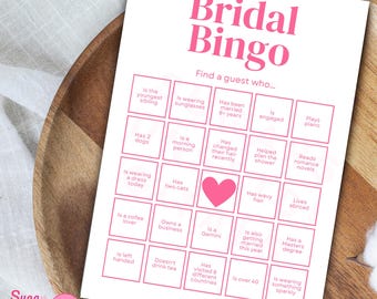 May include: A pink and white Bridal Bingo game card with a heart graphic. The card features a grid with prompts like "Is wearing sunglasses" and "Has two cats." The text "Bridal Bingo" is at the top, and the logo "Sugar Sunset Digitals" is at the bottom.
