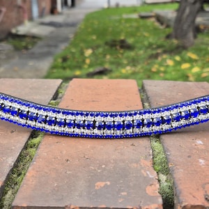 May include: A horse browband made of dark brown leather, featuring a curved design. The central section is adorned with rows of blue and clear crystals, creating a sparkling effect. This browband is designed to fit across the horse's forehead.