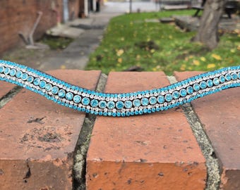 May include: A horse browband with a curved design. The browband is decorated with rows of light blue and clear crystals. The leather is a dark brown colour. The browband is resting on a red brick surface.