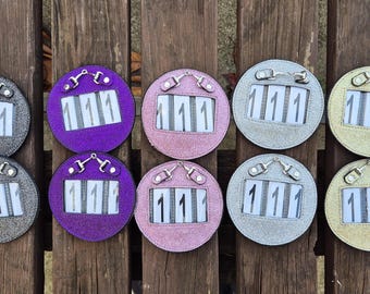 May include: Ten circular horse show number holders in shades of grey, purple, pink, silver, and gold. Each has the number "111" in white. The holders are displayed on a wooden surface.