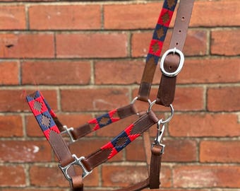 May include: A brown leather horse halter with red, blue, and brown patterned fabric accents. The halter features silver-colored metal hardware, including rings and a clasp. The background is a red brick wall.