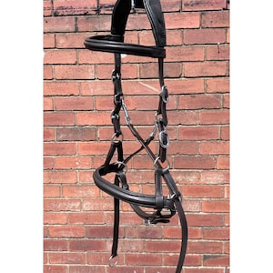 Op de afbeelding: Een zwart leren paardenhoofdstel met zilverkleurige metalen beslag. Het hoofdstel heeft een gewatteerde frontriem en neusriem en hangt voor een rode bakstenen muur. Ontworpen voor gebruik in de paardensport.
