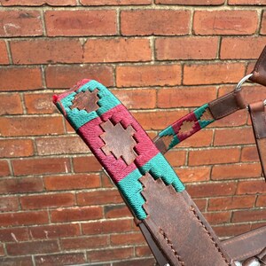 May include: A close-up of a brown leather horse bridle with teal and maroon woven detailing. The bridle has a decorative pattern and metal fittings. The background is a red brick wall.