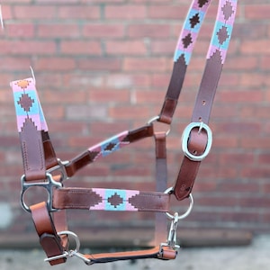 Pode incluir: Cabresto de cavalo em couro castanho com um padrão decorativo tecido em rosa, azul e castanho. O cabresto apresenta ferragens metálicas prateadas, incluindo fivelas e clipes. O padrão tecido é um desenho geométrico repetitivo.